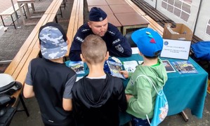 policjant siedzi przy biurku przed szkoła wokół 3 uczniów