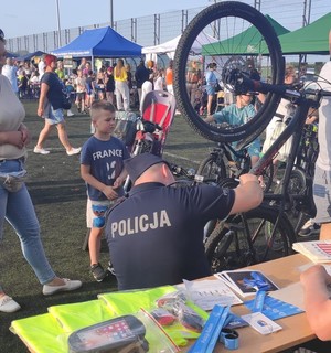 policjanta widac tyłem gdy znakuje rower na placu przed szkołą wokół grupa uczestników festynu