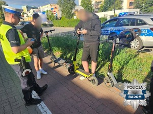 policjant konktrolujacy na ścieżce 3 kierujących hulajnogami, w tle radiowóz