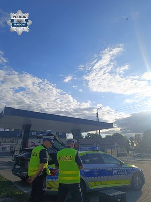 policjanci stoja tyłem do aparatu obok radiowóz, widac wznoszący się dron