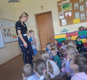 policjantka i chłopiec stoja na środku sali po prawej grupa dzieci siedzi na podłodze