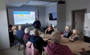 policjant na sali z grupą seniorów siedząca przy stoliku w tle ekran z prezentacją