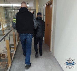 nieumundurowany policjant z opaska na ramieniu prowadzi mężczyznę obaj są tyłem do aparatu