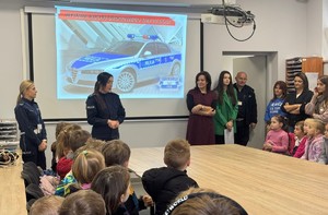 na sali dwie policjantki, grupa dzieci i ich opiekunki w tle ekran obraz radiowozu