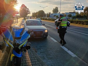 policjant podchodzi do czarnego auta, które zatrzymuje do kontroli, po lewej stronie widac policyjny motocykl
