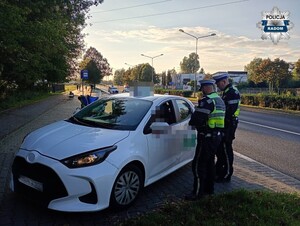 dwaj policjanci  z drogówki stoi przy białym aucie osobowym, kontroluja kierowcę, a  w tle widac policyjne motocykle