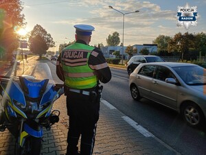 policjant stoi tyłem obserwuje ruch po jego lewej stronie motocykl w tle auta na ulicy