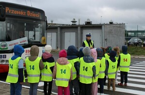 policjantka i grupa przedszkolaków w kamizelkach odblaskowych obok widac autobus komunikacji miejskiej