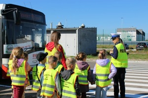 policjantka i grupa przedszkolaków w kamizelkach odblaskowych obok widac autobus komunikacji miejskiej