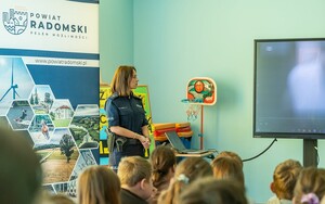 policjantka stoi na sali na spotkaniu w tle ekran widac głowy siedzących uczniów