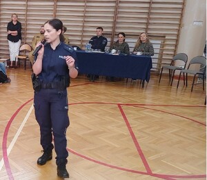 policjantka na srodku sali trzyma mikrofon w tle przy stoliku siedza przedstawiciele innych służb