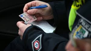 zdjęcie przedstawia ręce, które w dłoniach trzymają blankiet prawa jazdy oraz długopis, na jednym z rękawów widoczna jest naszywka z literą &quot;R&quot;. Na zdjęciu widoczna jest również fragment odblaskowej kamizelki z napisem &quot;Policja&quot;