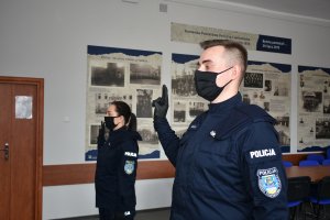 zdjęcie przedstawia kobietę i mężczyznę w granatowym policyjnym mundurze, którzy stoją koło siebie trzymając w górze rękę ze złączonymi dwoma palcami