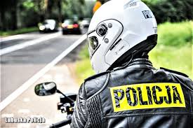 grafika przedstawia osobę ubraną w biały kas, czarna skórzana kurtkę z żółtym napisem na plecach &quot;Policja&quot;