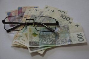 Fotografia kolorowa: na białym tle banknoty w walucie polskiej na których widzimy okulary w czarnych oprawkach.