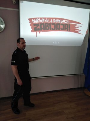 Fotografia kolorowa: na tle białej tablicy z napisem kampanii &quot;Narkotyki i dopalacze zabijają&quot; widzimy dzielnicowego z KPP Mława.