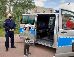 umundurowany policjant stojący przy radiowozie pozuje do zdjęcia z dzieckiem ubranym w kask policyjny