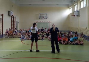 dwie umundurowane policjantki stojące w sali gimnastycznej przed grupą siedzących dzieci