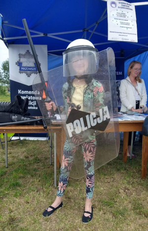 dziewczynka ubrana w sprzęt przeciwuderzeniowy stojąca przy niebieskim namiocie Policji