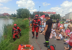 strażacy ubrani w sprzęt do ratownictwa wodnego stojący przed grupą dzieci