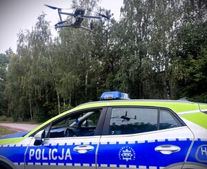 Na zdjęciu widać policyjny radiowóz stojący na tle wysokich drzew. Samochód jest oznakowany — na jego boku znajduje się duży napis „POLICJA”. Karoseria ma charakterystyczne niebiesko‑żółte elementy odblaskowe, a na dachu zamontowana jest niebieska belka świetlna.

Radiowóz stoi na utwardzonej nawierzchni, prawdopodobnie przy drodze lub na parkingu w pobliżu lasu. W tle widać gęste, wysokie drzewa, które zajmują całą górną część kadru.

Nad radiowozem, na wysokości kilku metrów, unosi się dron policyjny. Jest wyraźnie widoczny na tle nieba. Ma cztery ramiona z wirnikami, typowe dla dronów wykorzystywanych do monitoringu. W szybie radiowozu odbija się jego sylwetka — widać zarówno drona w powietrzu, jak i jego odbicie w bocznej szybie pojazdu.