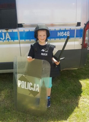 Dziecko na tle radiowozu policyjnego wyposażone w sprzęt PZ. Chłopiec ma na sobie kamizelkę ochronną, na głowie specjalny kask ochronny, w ręku trzyma pałkę szturmową i tarczę.