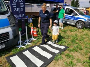 Policjantka udzielająca dziecku wskazówek jak prawidłowo korzystać z dróg. Funkcjonariuszka przeprowadza chłopca po macie symulującej przejście dla pieszych.