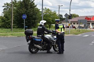 Policjant siedzący na motocyklu rozmawia z policjantem stojącym na środku skrzyżowania