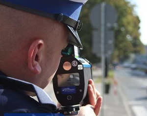 Zdjęcie przedstawia policjanta patrzącego przez urządzenie do pomiaru prędkości