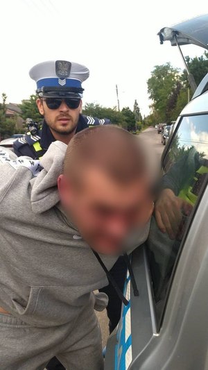 Policjant w okularach trzyma za ręce trzymane z tyłu mężczyznę z siwym dresie