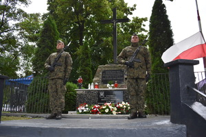 Pomnik z krzyżem. Przed pomnikiem stoi dwóch żołnierzy. Z lewej strony widać flagę biało-czerwoną