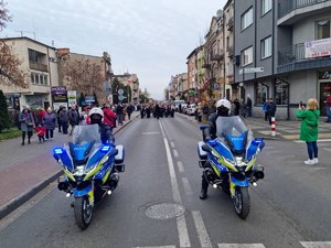 Dwaj policjanci jadą na motocyklach. Za nimi idą uczestnicy uroczystości