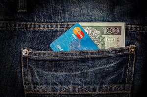 Kieszeń granatowych spodni dżinsowych z których wystaje karta płatnicza i banknot 100 złotych