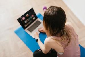 Kobieta siedząca na niebieskiej macie do ćwiczeń patrzy w kierunku laptopa, który stoi przed nią