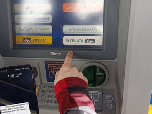 Zdjęcie przedstawia wyświetlacz bankomatu, fragment dłoni i wyświetlacz telefonu komórkowego z wiadomością o przesłanych środkach.