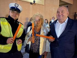 Kobieta i mężczyzna stoją obok policjanta. Trzymają w ręce pomarańczowe odblaski