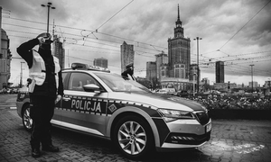 Czarno-białe zdjęcie przedstawiające policjanta salutującego, stojącego przy radiowozie. W tle Warszawa
