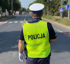 Policjant w białej czapce i odblaskowej kamizelce stoi tyłem i patrzy w kierunku drogi