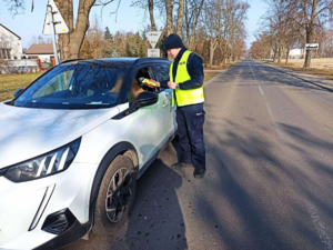 Policjant stoi przy białym samochodzie i trzyma sprzęt służący do badania stanu trzeźwości kierowców