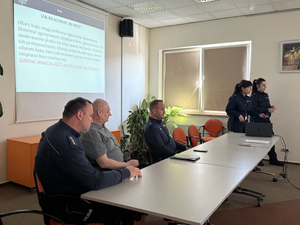 Policjanci siedzą przy stole i stoją obok ściany.