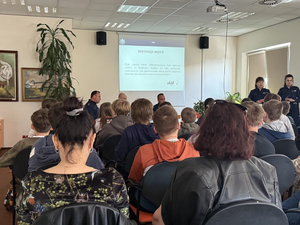 Policjanci siedzą przy stole i stoją obok ściany. Przed nimi siedzą uczestnicy debaty.