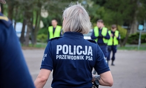 Tyłem stoi kobieta w granatowej koszulce z napisem policja przewodnik psa. w tle widać policjantów ubranych w kamizelki odblaskowe.