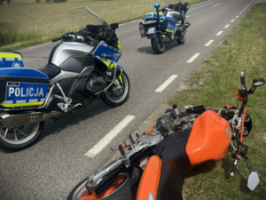 Pomarańczowy motocykl leży na ziemi. Dwa policyjne motocykle stoją na drodze