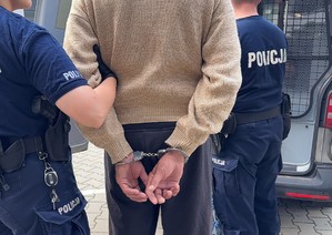 Policjantka trzyma za rękę mężczyznę skutymi kajdankami. W drugim planie widać policjanta przy radiowozie