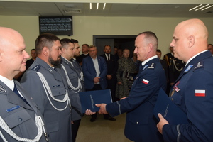 Dwóch policjantów stoi naprzeciwko szeregu policjantów. Jeden z nich podaje teczkę policjantowi stojącemu w szeregu