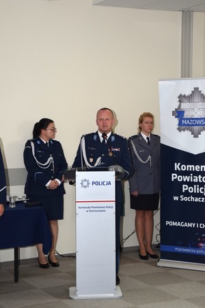 Policjant stoi przy mównicy. Za nim stoją policjantki
