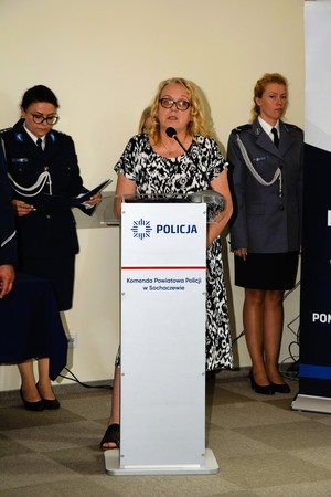 Kobieta stoi przy mównicy, a za nią stoją dwie policjantki