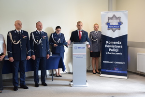Mężczyzna stoi przy mównicy, a za nim stoją dwie policjantki. Obok stoi dwóch policjantów