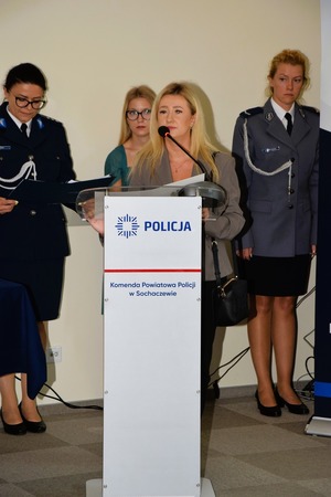 Kobieta stoi przy mównicy, a za nią stoją dwie policjantki i pracownik policji