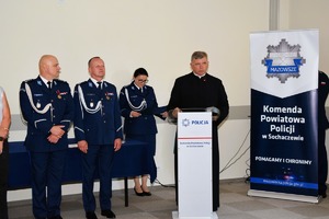 Ksiądz stoi przy mównicy, a za nim stoją dwie policjantki